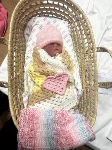 Wlsie in a moses basket wearing a pink hat, rapped in a pink, yellow and beige crocheted blanket with a crocheted heart resting on top.