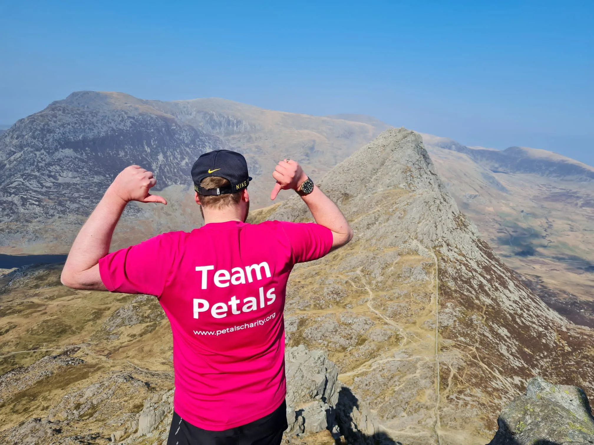 Guy on Everest, back to camera with his pink Petals t shirt on, thumbs pointing back to the white Team Petals wording on his back