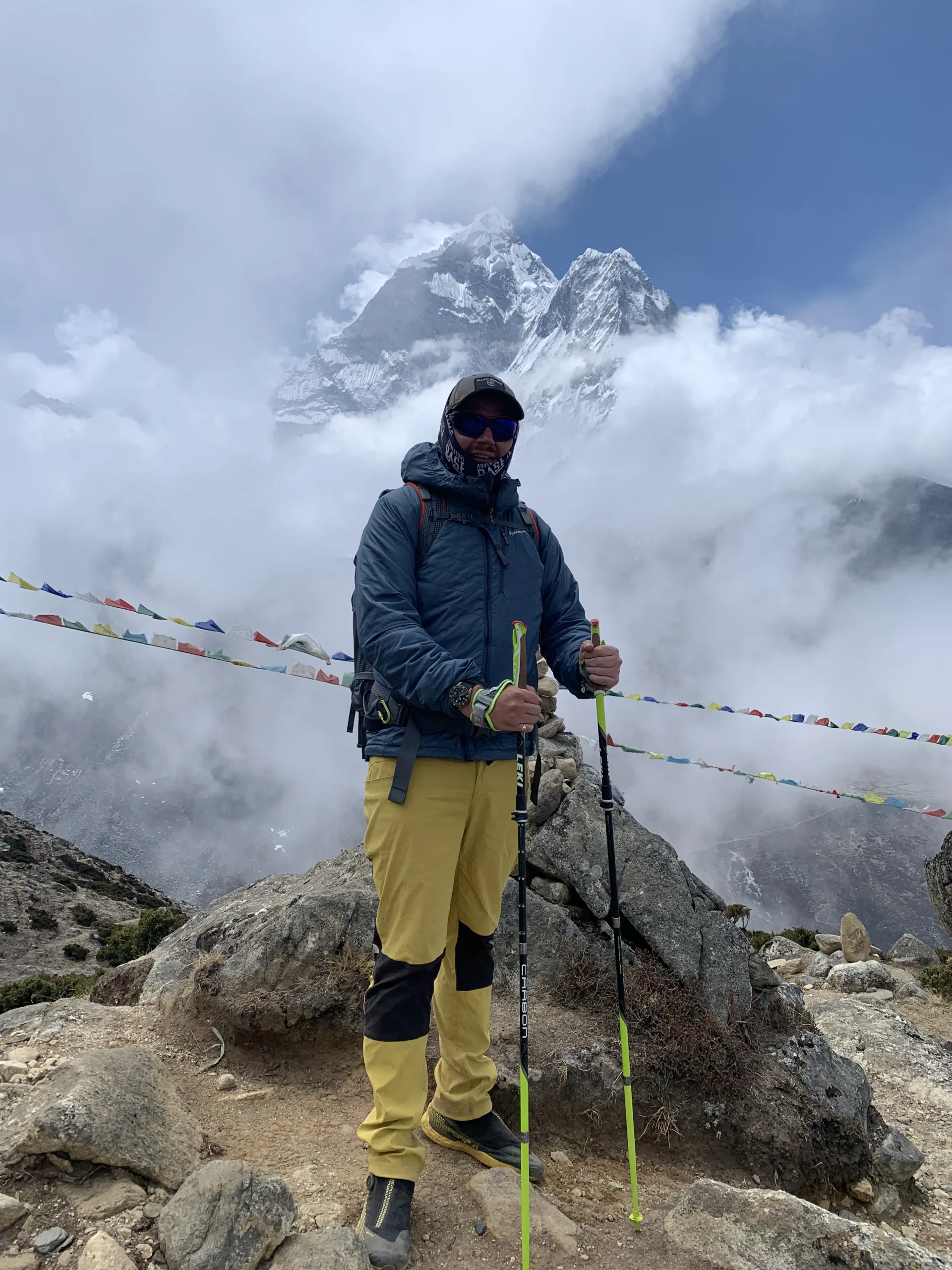 Guy on Everest, mists and mountains in background, holding walking sticks, looking at camera.