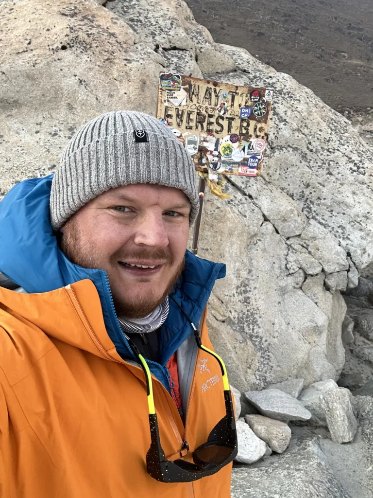Guy in orange waterproof smiling to camera with rocks and Everest sign behind him
