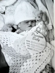 Elsie in black and white surrounded by teddies, wrapped in a crocheted blanket and with a Precious baby tag bearing her name - Elsie Jacqueline'.