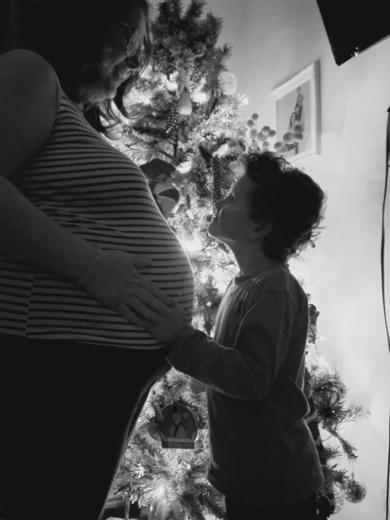Rebecca and Ollie standing opposite each other in front of a Christmas tree, Ollie is cradling her bump.